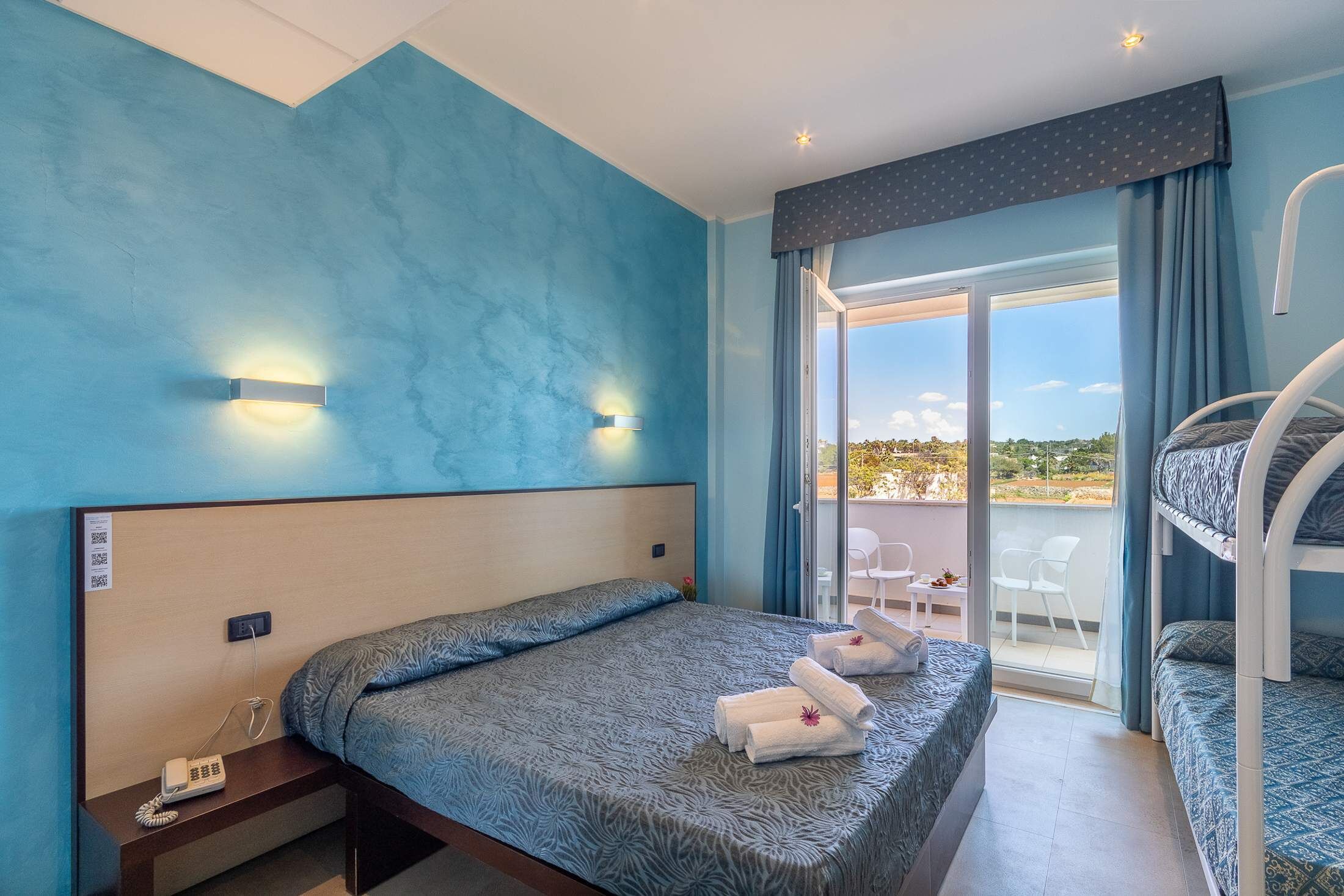 Ein Schlafzimmer mit Bett, Nachttischtelefon, Balkon mit Stühlen und Blick ins Freie.