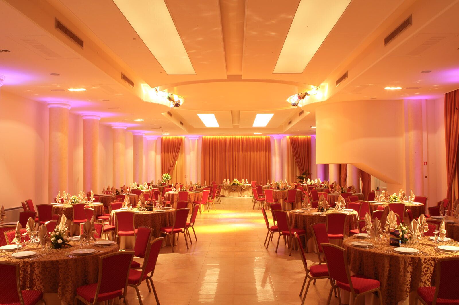 A banquet hall with round tables and red chairs, arranged in rows.