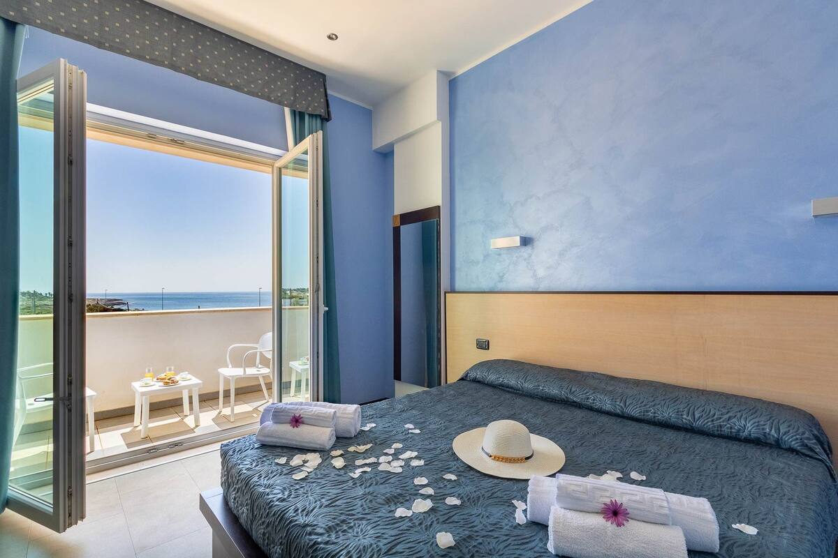 A hotel room with a large bed, a balcony with a view, and a small table with a chair.