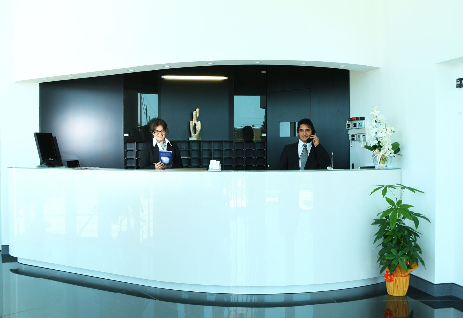 A reception desk with two staff members, a laptop, a vase with flowers, and a potted plant.