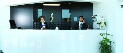 A reception desk with two staff members, a laptop, a vase with flowers, and a potted plant.
