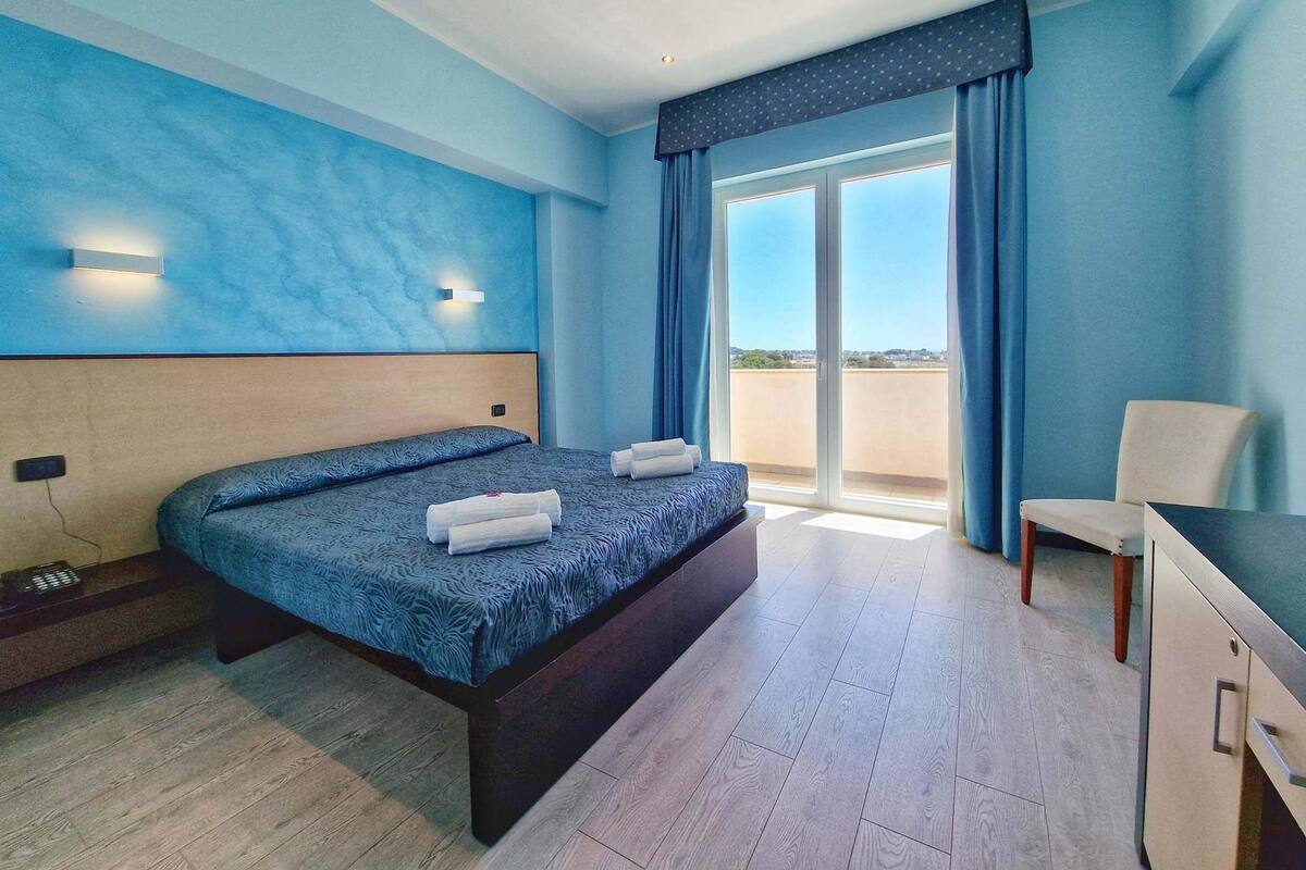 A modern hotel room with a large bed, a desk, and a view of the outdoors through a sliding glass door.