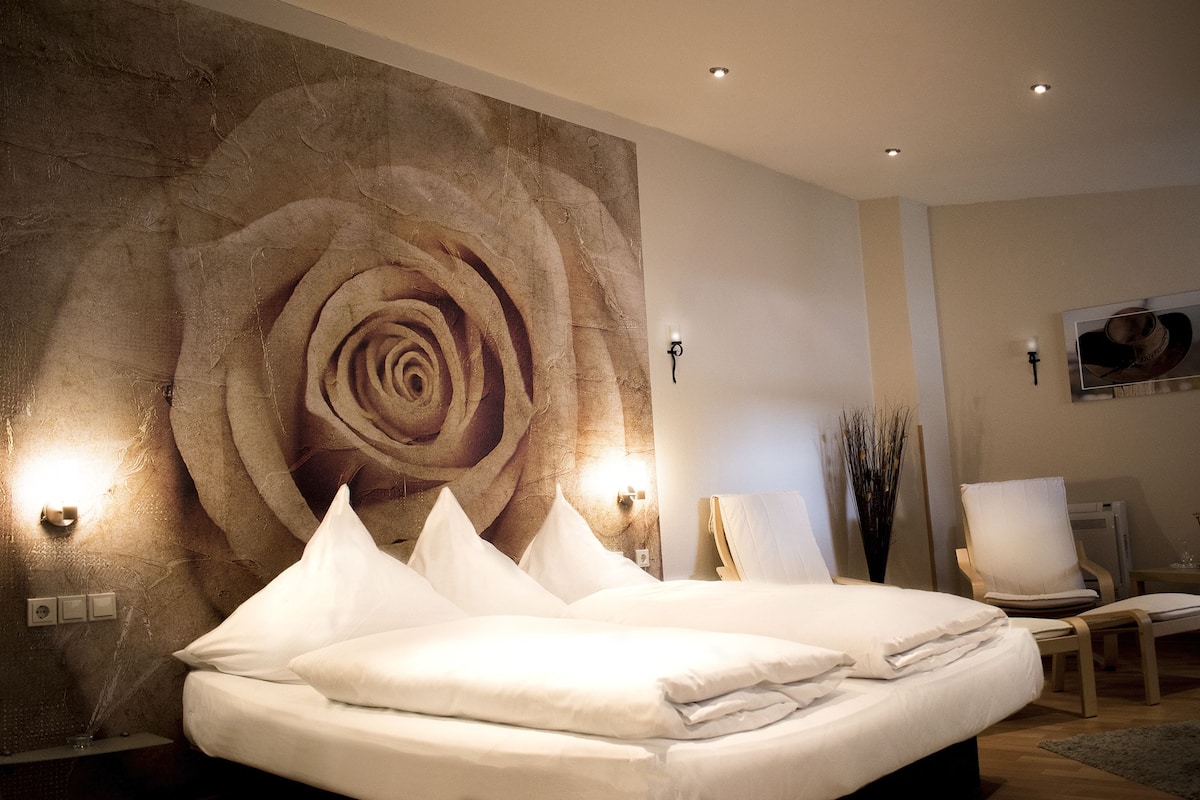 A bedroom with a large wall mural of a rose, a bed with white linens, a chair, and a small table.
