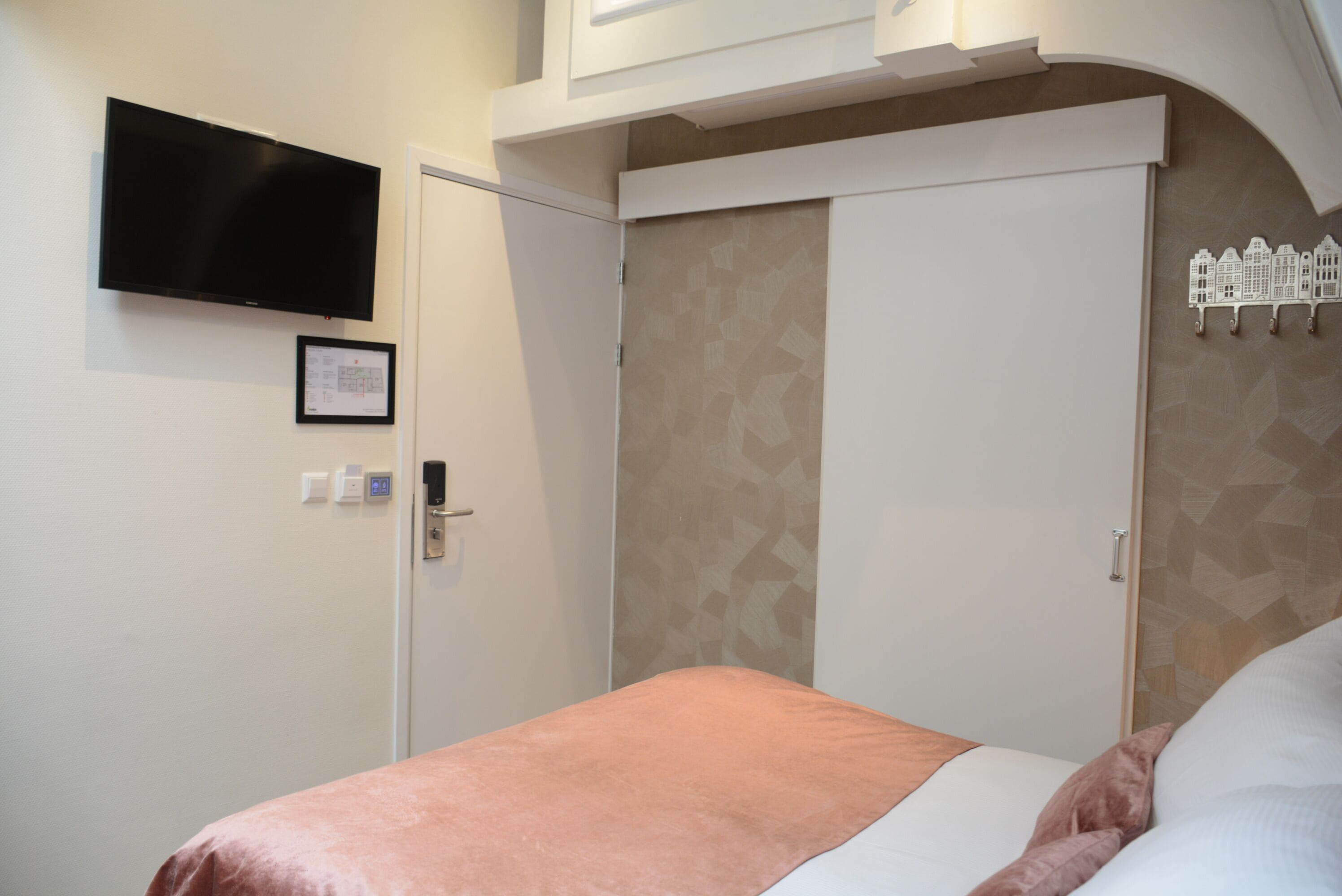 A hotel room with a bed, a flat-screen TV mounted on the wall, a door with a handle, and a wall-mounted light switch.