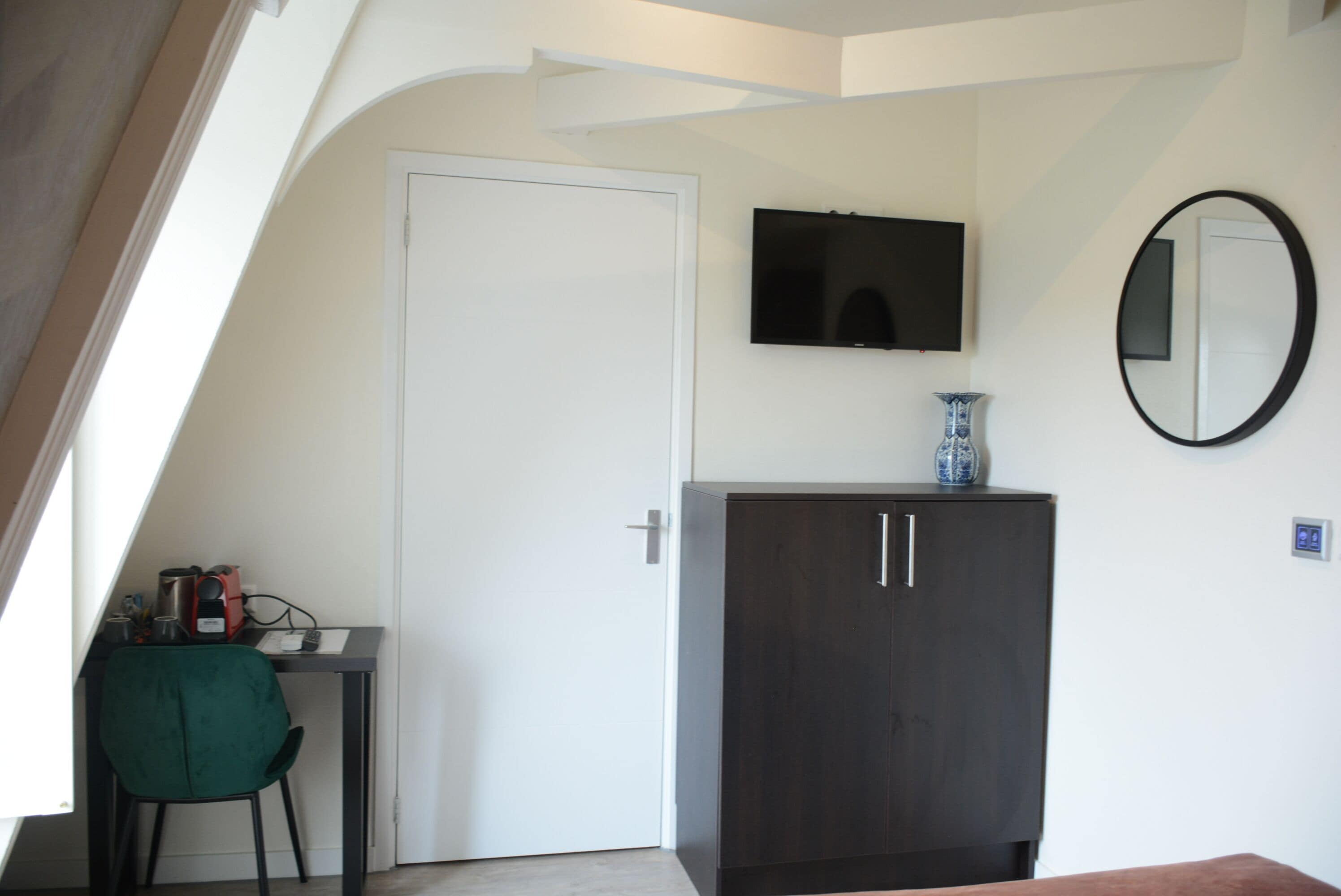 A room with a white door, a dark wooden cabinet, a small table with a green chair, a flat-screen TV mounted on the wall, and a round mirror.