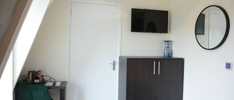 Una habitación con una puerta blanca, un mueble de madera oscura, una pequeña mesa con una silla verde, un televisor de pantalla plana montado en la pared y un espejo redondo.