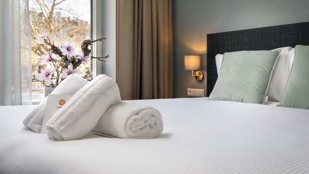 Una cama bien hecha con sábanas blancas y toallas dobladas, un florero con flores y una lámpara en la mesita de noche en una habitación de hotel.