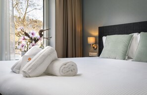 Un lit bien fait, avec du linge de lit blanc et des serviettes pliées, un vase rempli de fleurs et une lampe sur la table de chevet, dans une chambre d’hôtel.
