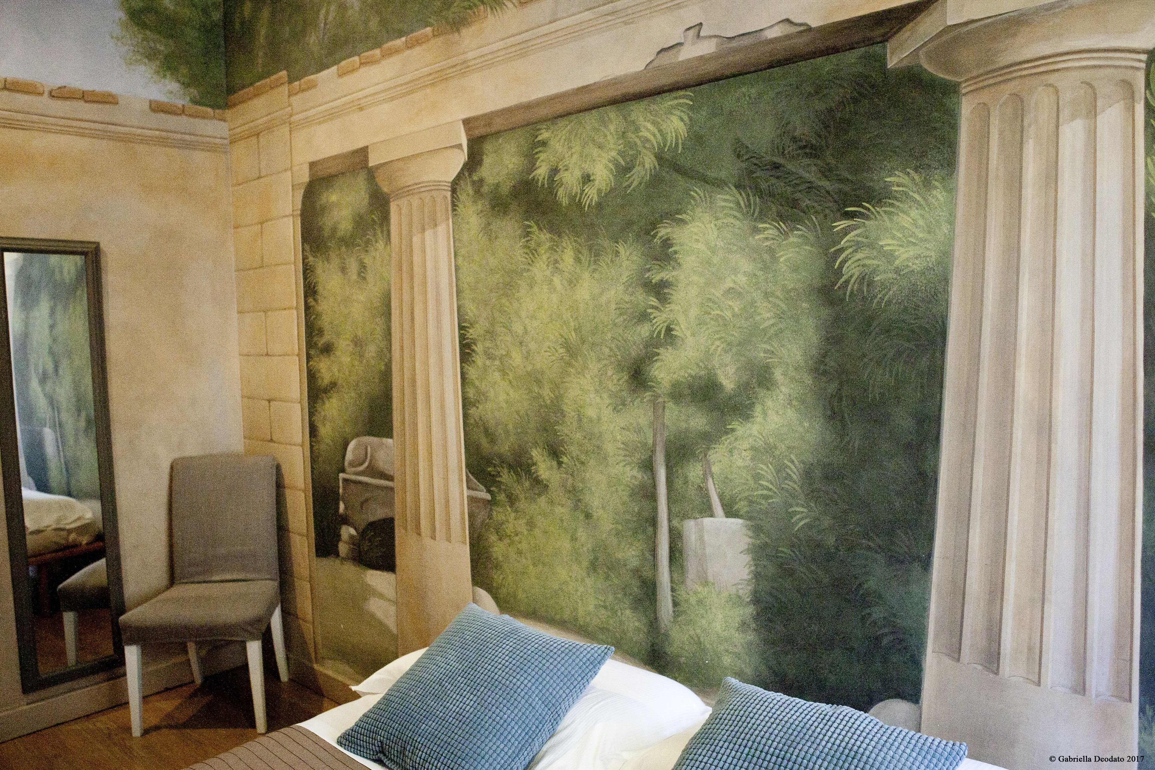 A room with a bed, a chair, and a mural of columns and foliage.