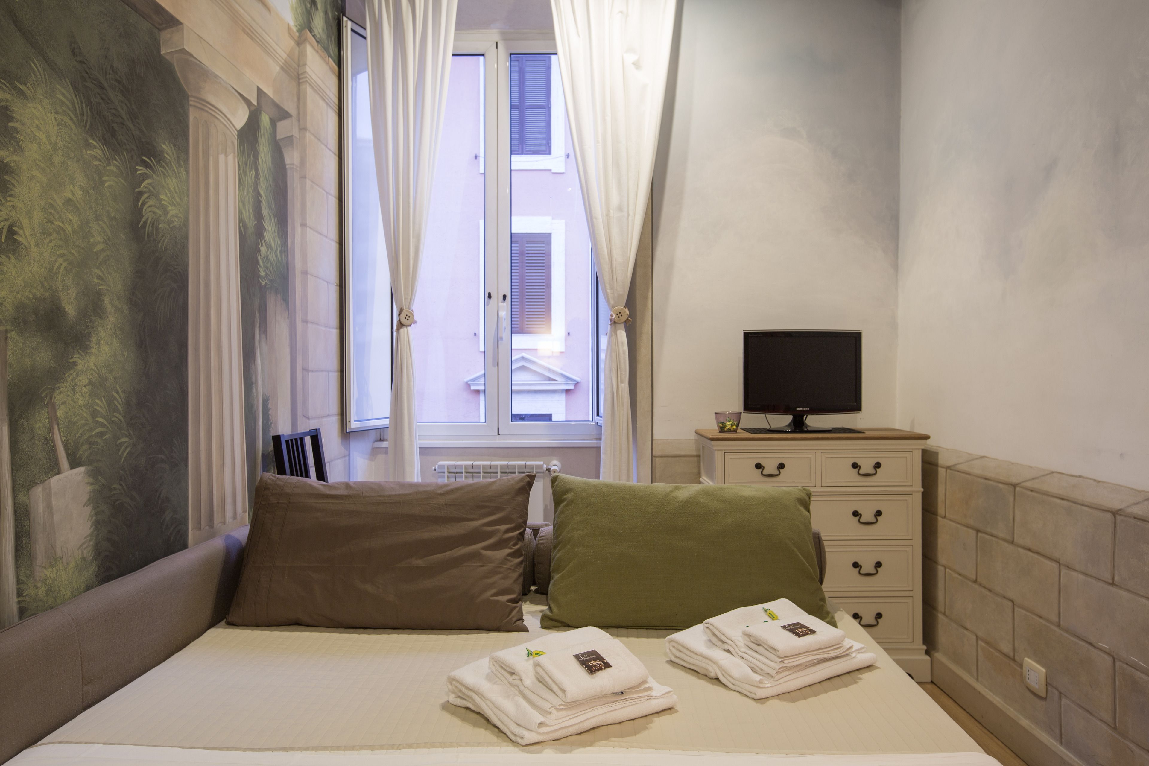 A hotel room with a bed, pillows, a TV on a dresser, and a window with curtains.