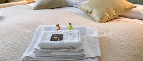 A neatly made bed with folded white towels and a small card, a beige pillow, and a view of greenery through a window.