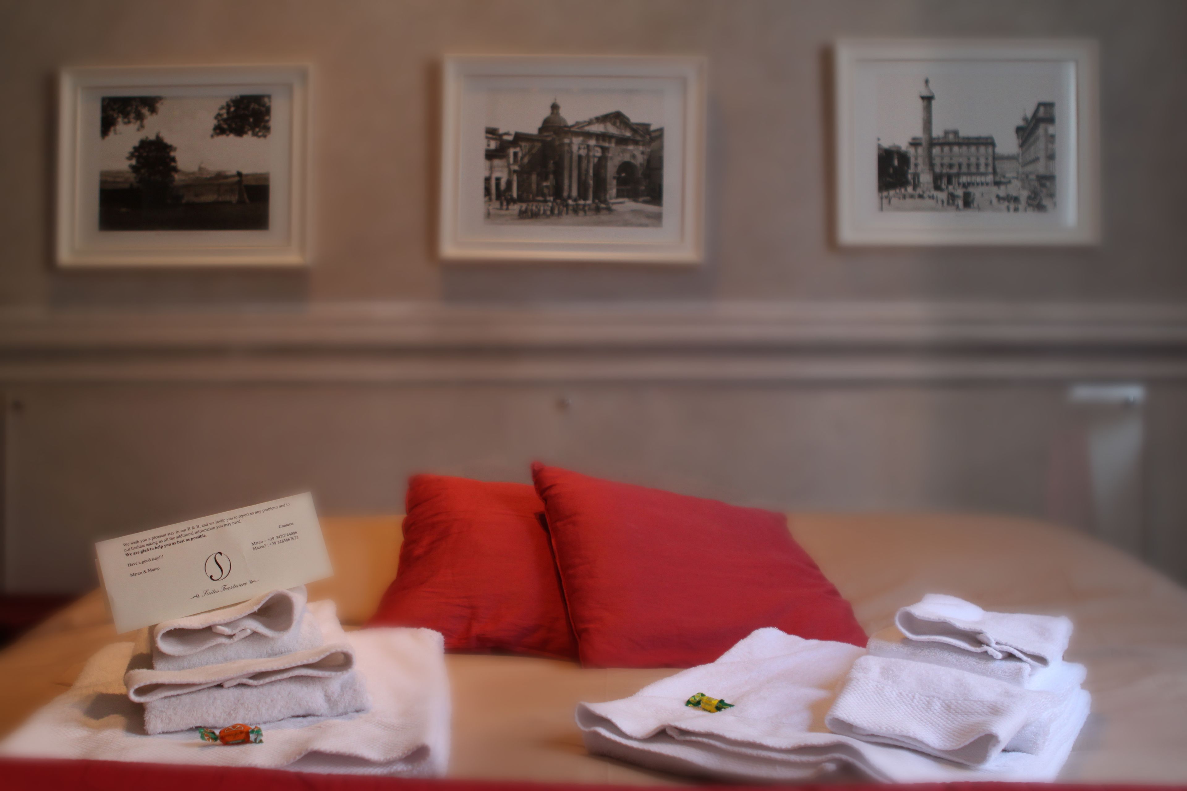 A neatly made bed with red pillows, a folded towel, and a card on the bed. There are three framed pictures on the wall above the bed.