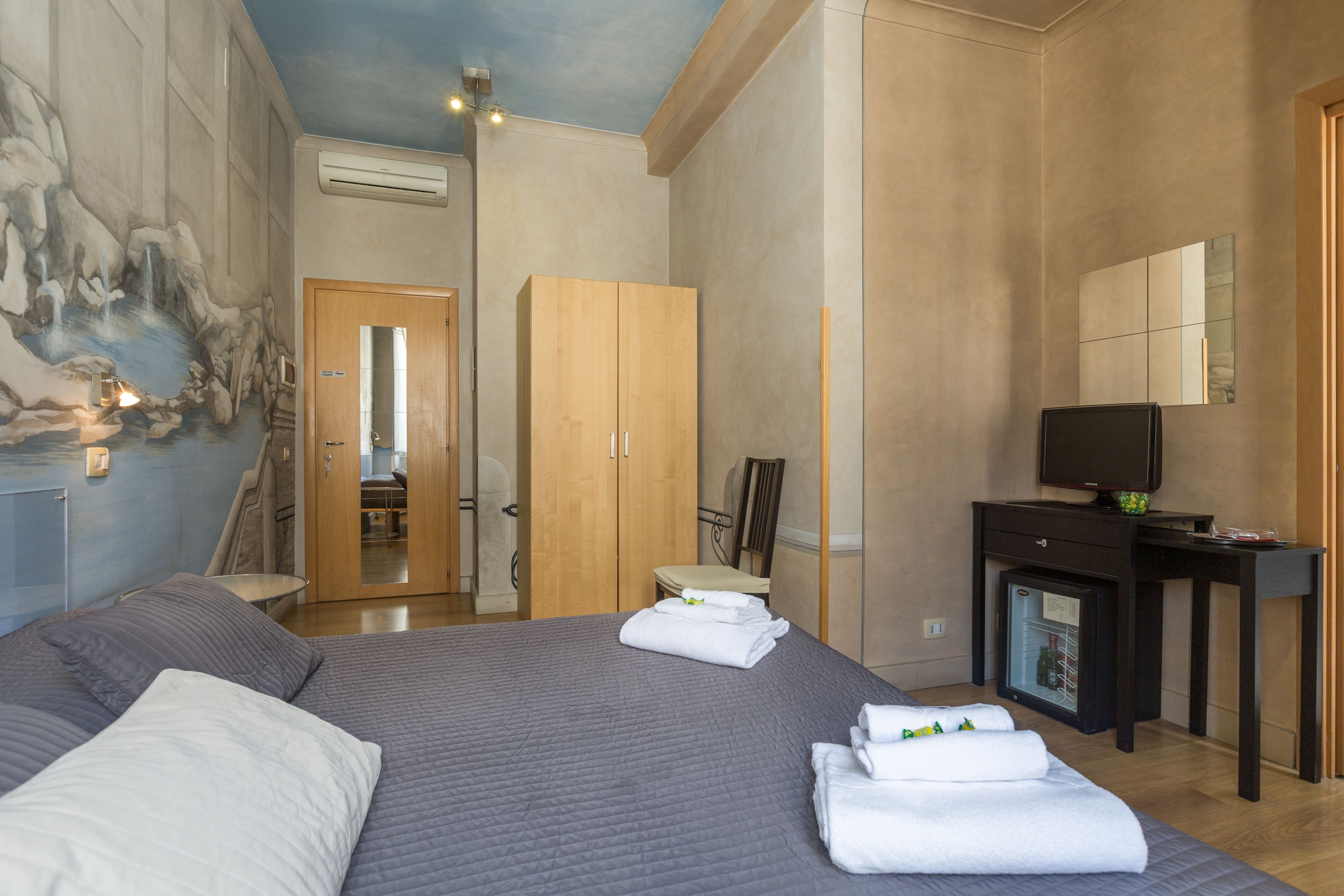 Une chambre d’hôtel avec un grand lit, un petit bureau avec une télévision, un minibar et une salle de bain visible à travers une porte ouverte.