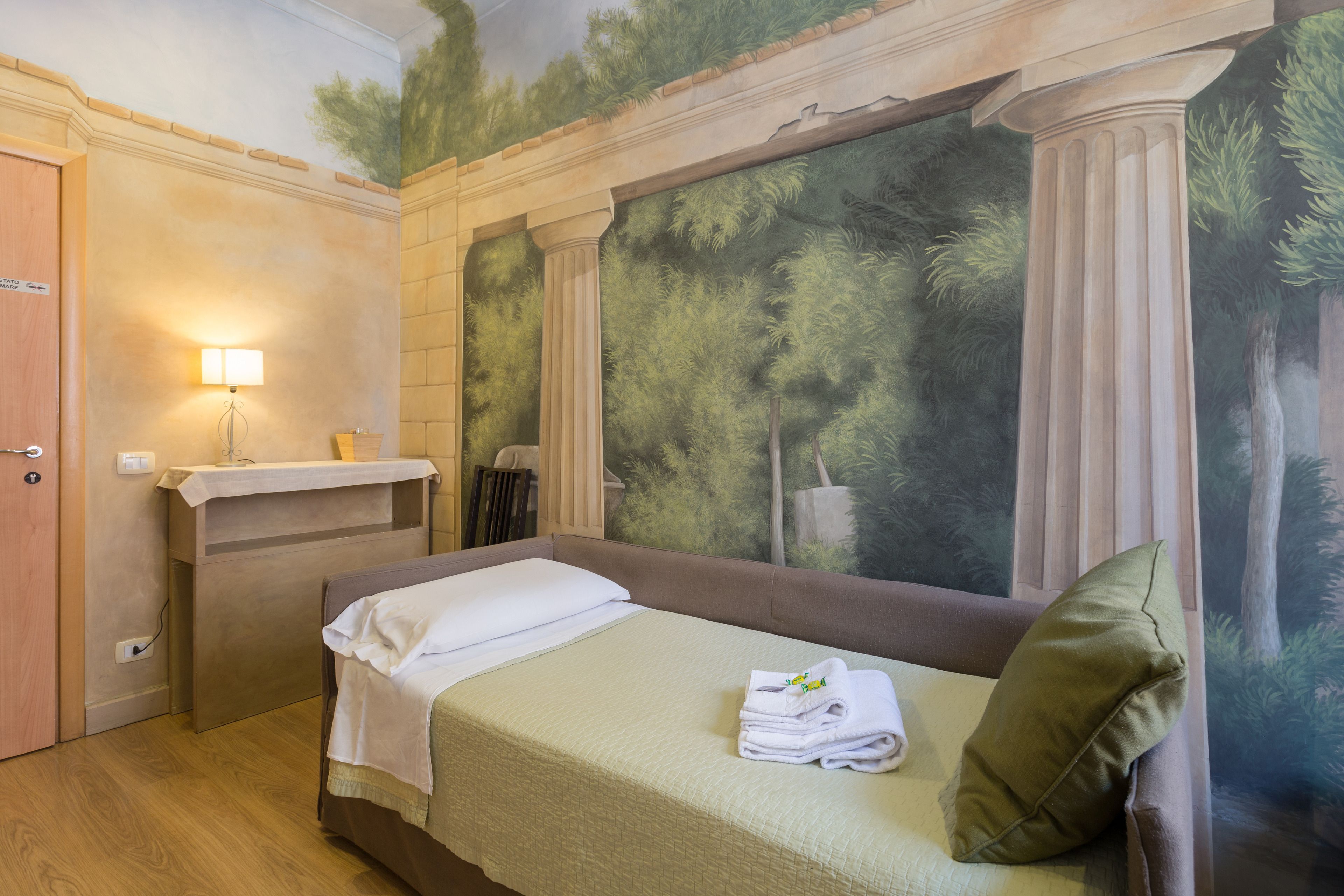 A hotel room with a bed, a nightstand, a lamp, and a mural of trees on the wall.