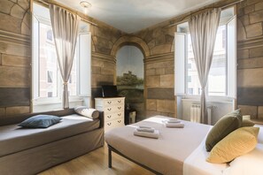 A bedroom with a stone wall, a bed with pillows and folded towels, a sofa with a pillow, and a dresser. - Suites Trastevere (Rome)