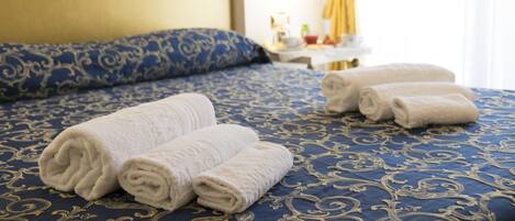 A neatly made bed with blue and gold patterned bedding, two rolled white towels, and a bedside table with a lamp and a small decorative item.
