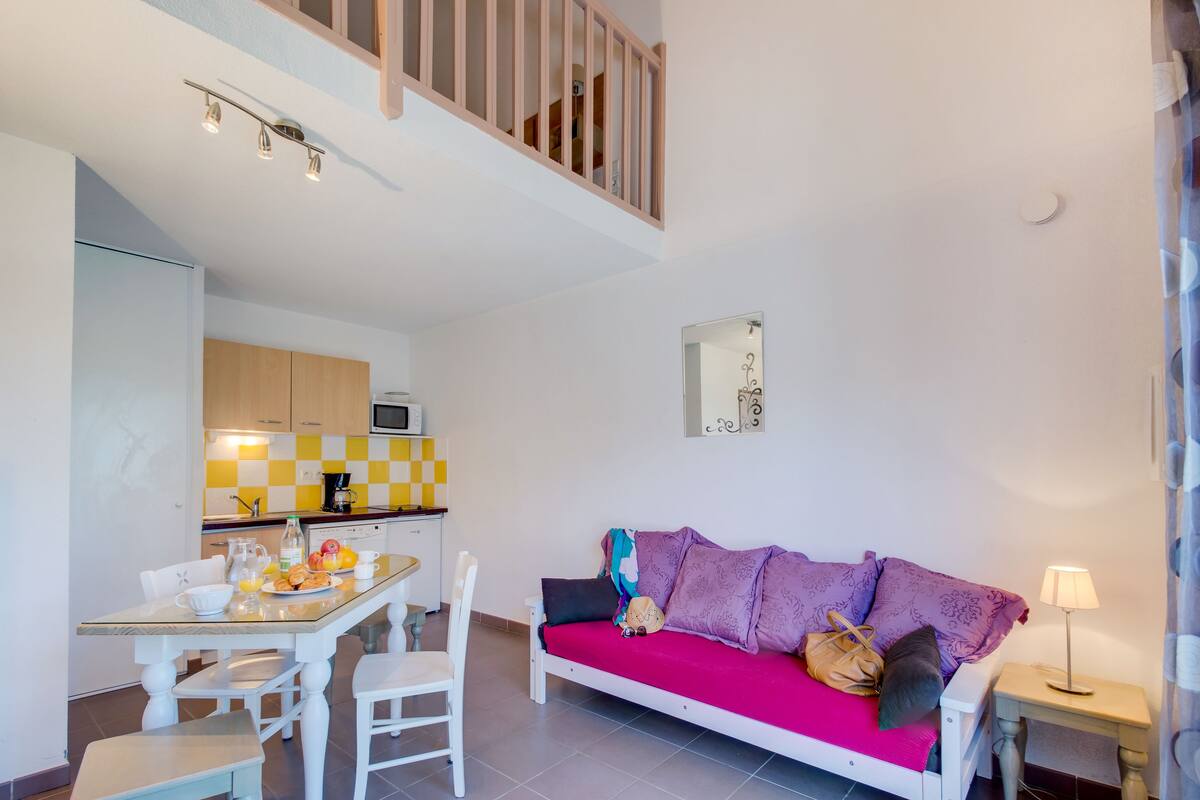A modern living room with a sofa, a dining table with chairs, a kitchen in the background, and a staircase.