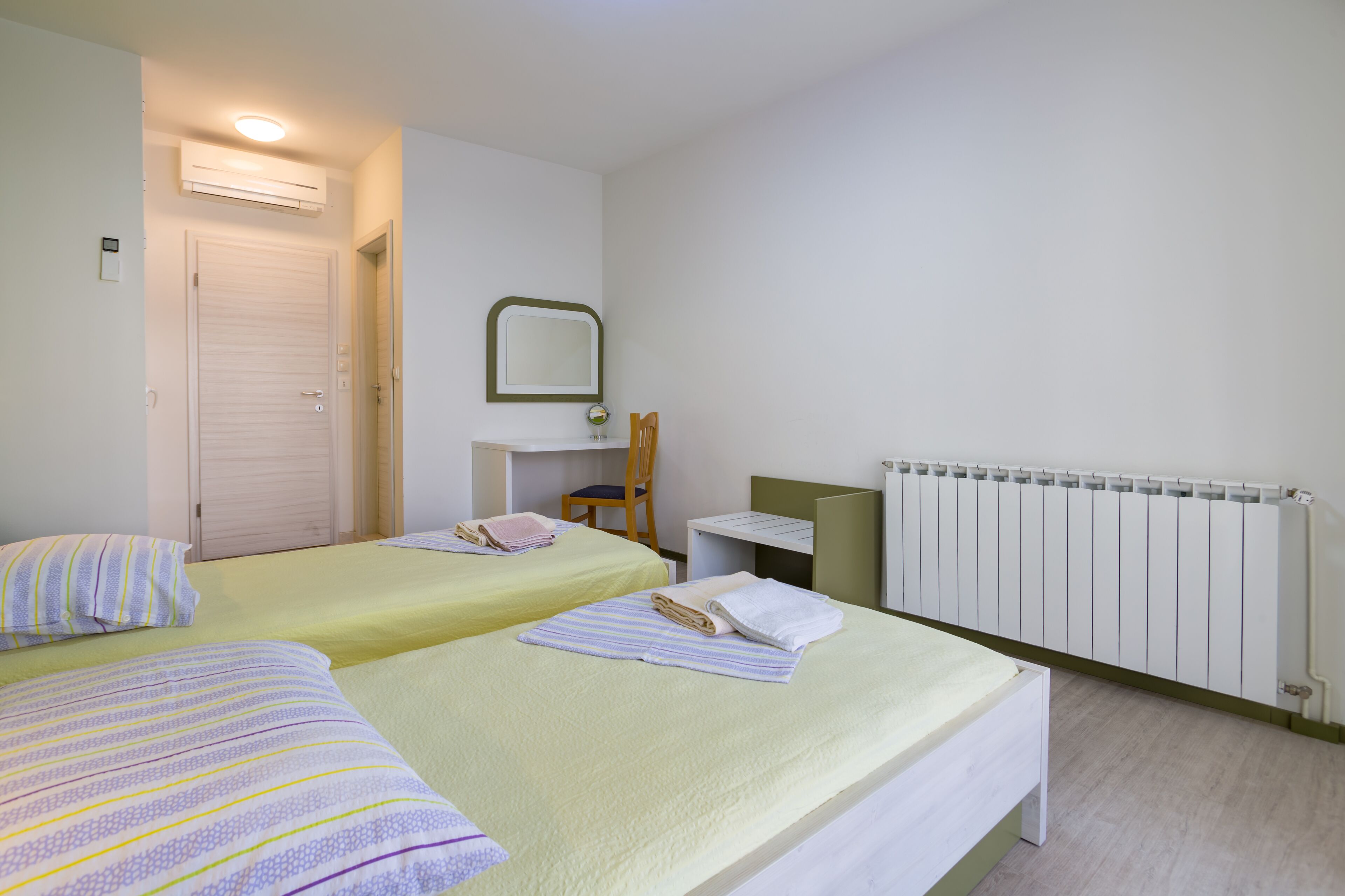 A hotel room with two beds, a chair, a desk, a mirror, and a radiator.