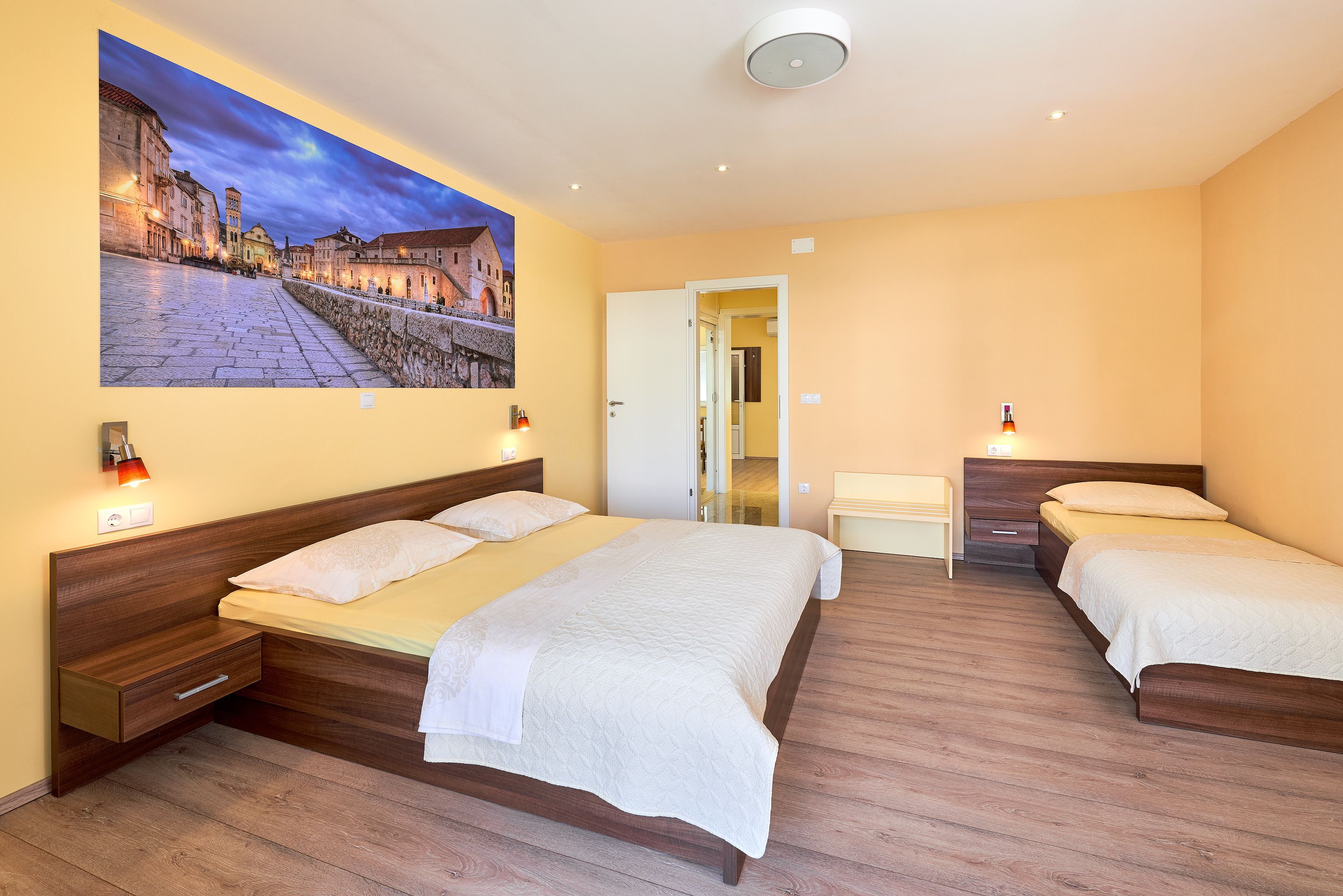 Una habitación de hotel con una cama grande, una cama más pequeña, una mesita de noche y una lámpara fijada a la pared.