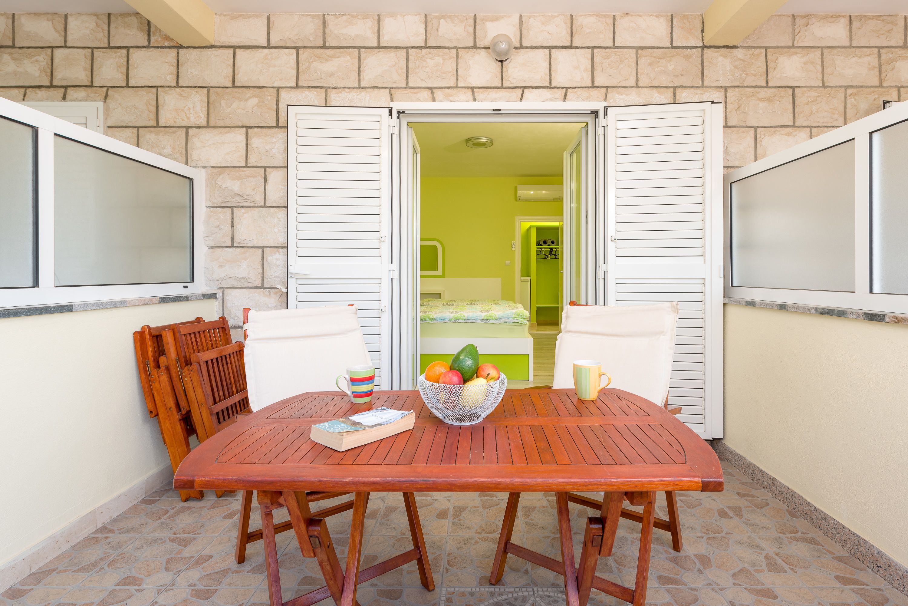 Una mesa de madera con sillas, un frutero y un libro en un balcón con suelo de piedra, contraventanas blancas y vistas a una habitación con cama y pared verde.