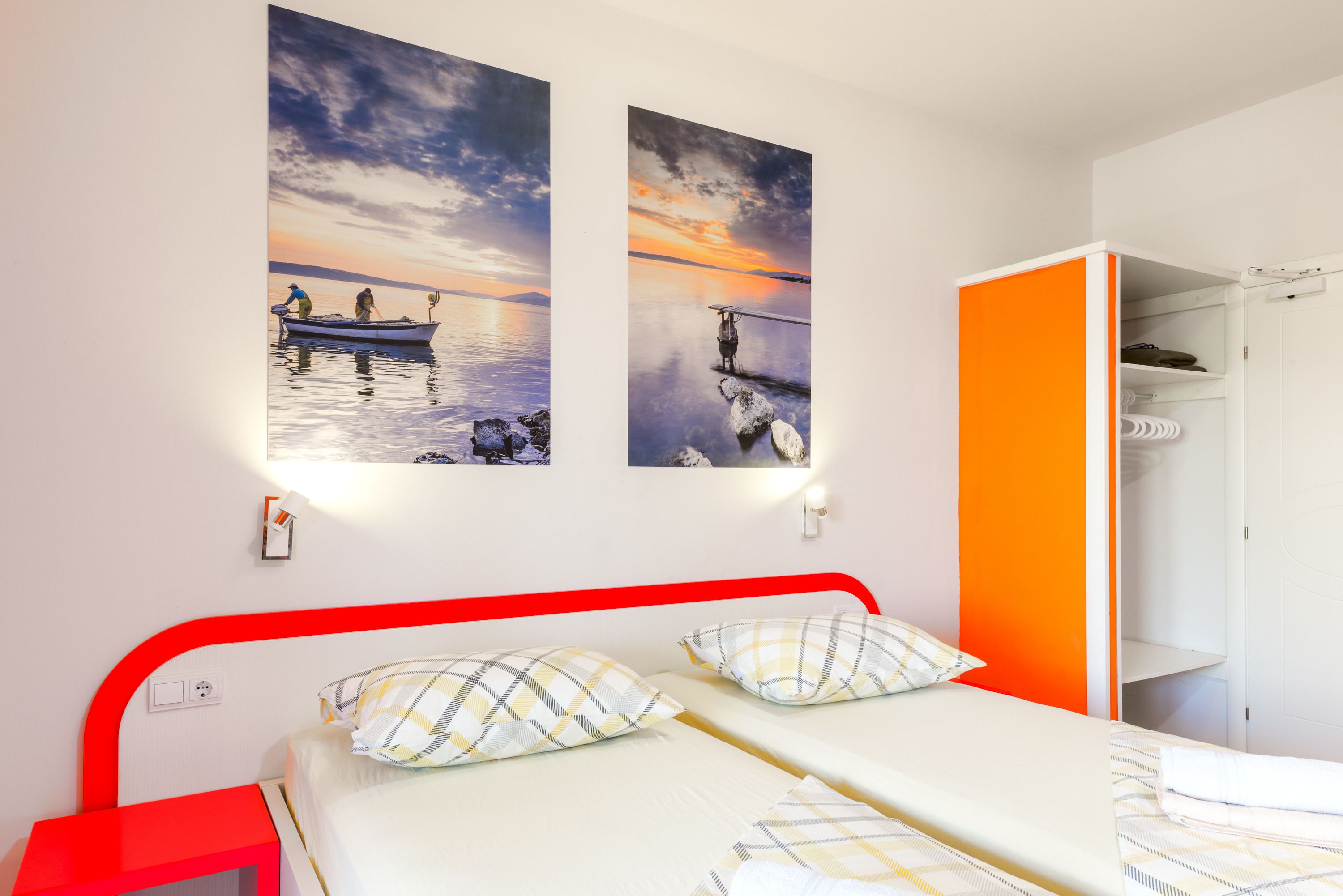 A double bed with two pillows, a red headboard, and a white nightstand. There are two wall-mounted pictures depicting a sunset over water. An orange closet is partially open to the right.