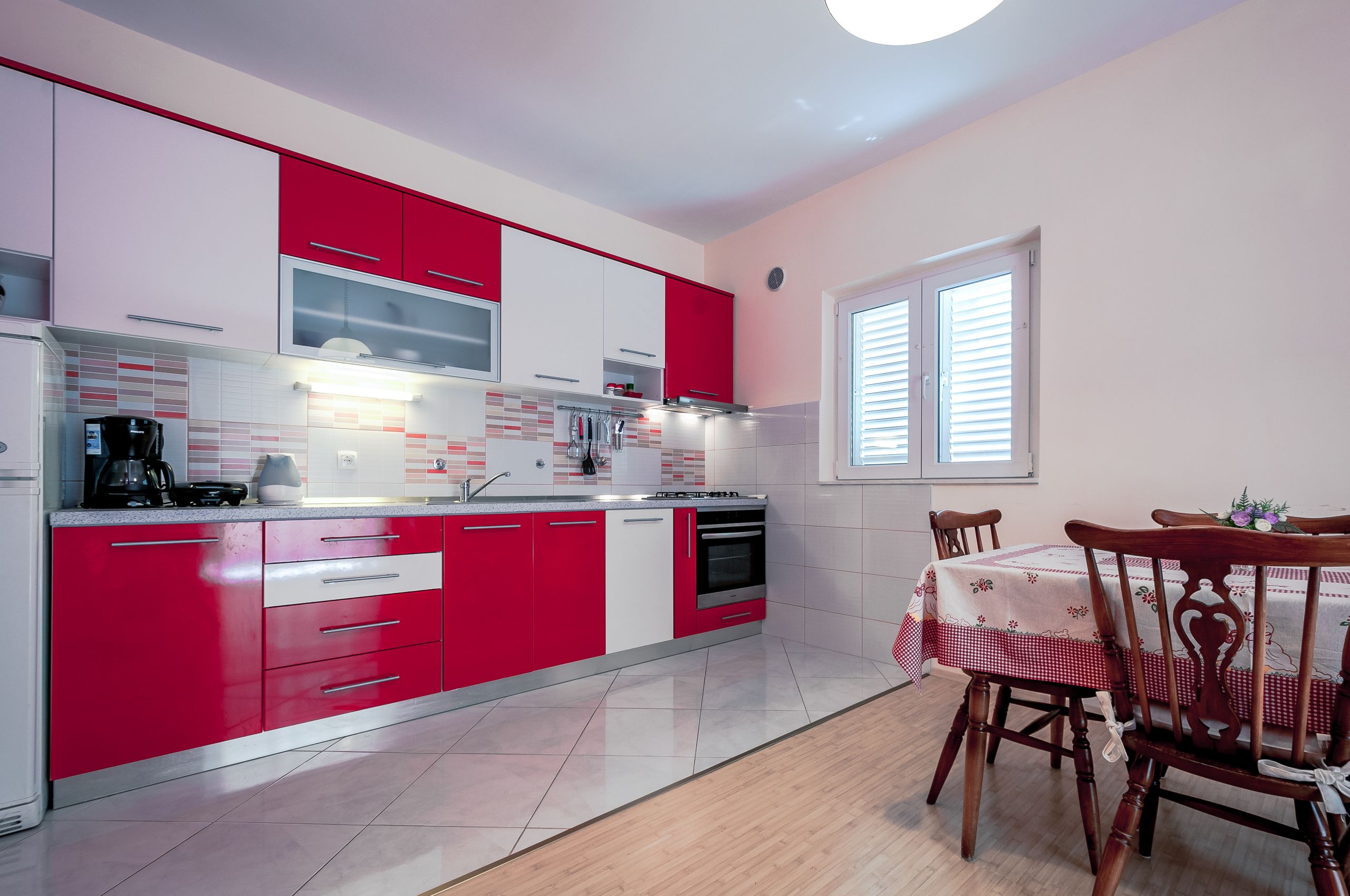 Une cuisine moderne avec des meubles de rangement rouges et blancs, un coin repas avec une table dressée pour deux personnes, et une fenêtre équipée de stores.