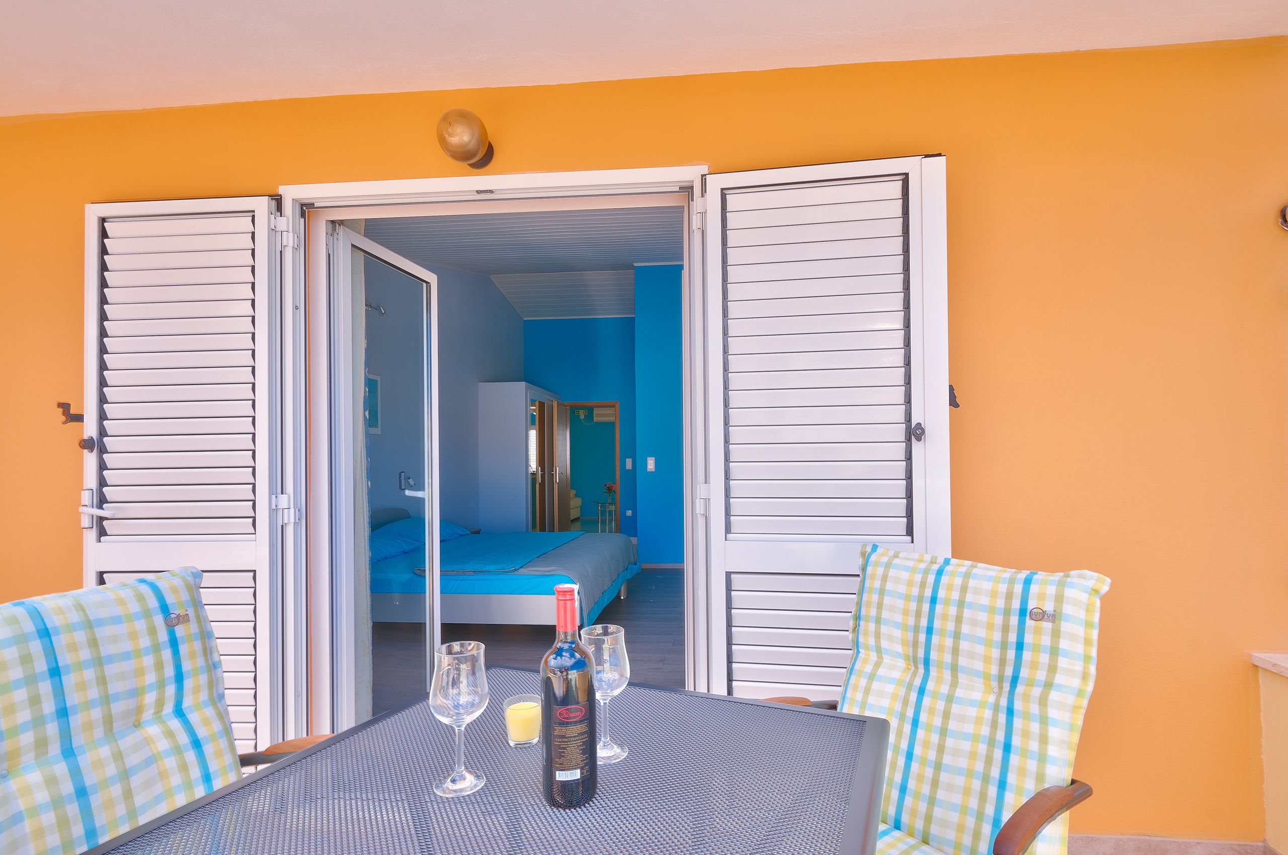 An open room with a bed, a table with a bottle and glasses, and checkered chairs.