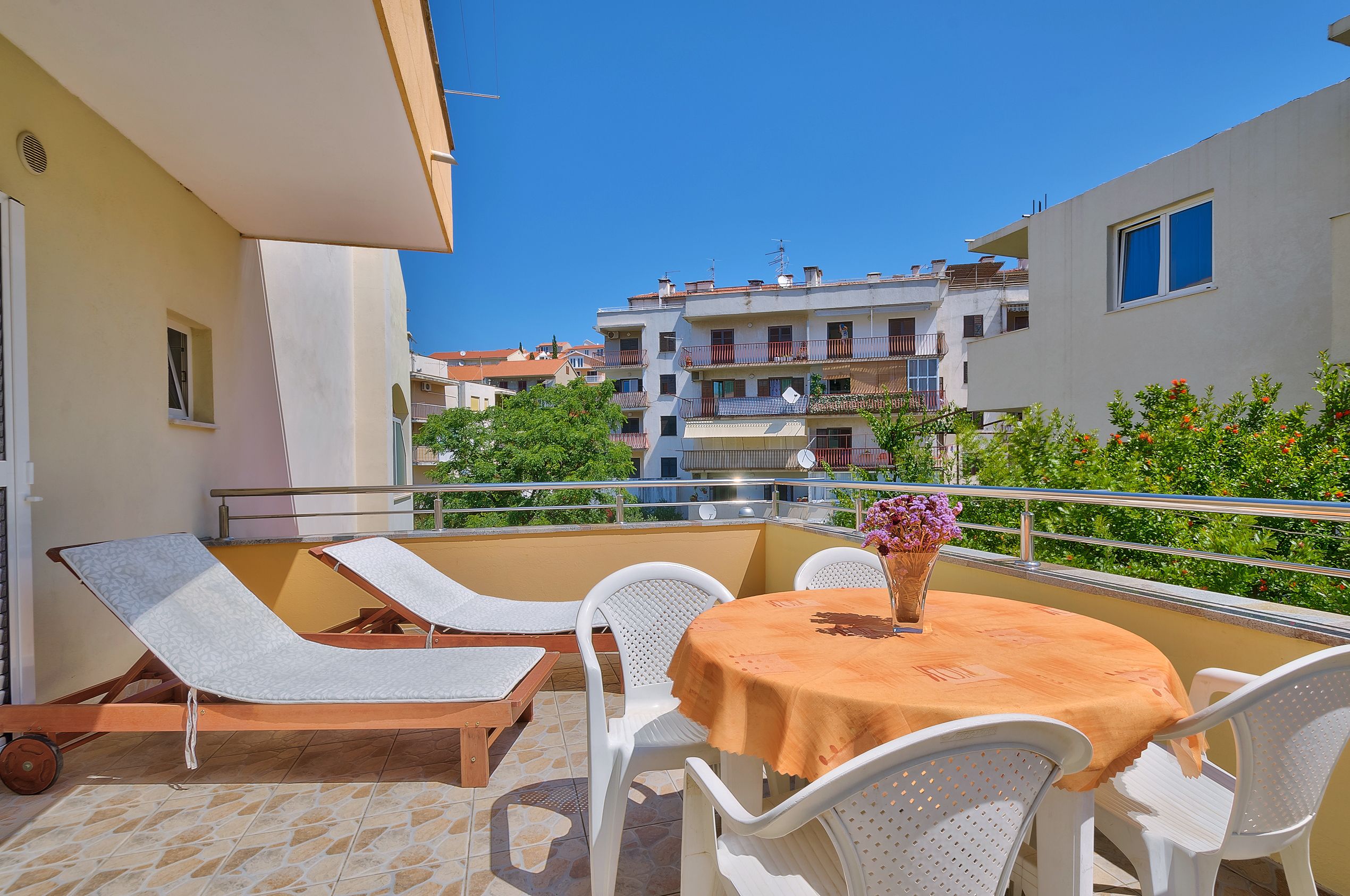 A balcony with a table and chairs, a sun lounger, and a view of neighboring buildings.