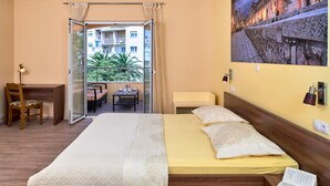 A hotel room with a bed, a desk, a chair, a small table, and a balcony with a view of buildings and greenery.