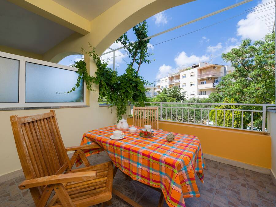 Un balcon avec une chaise et une table en bois, recouverts d’une nappe colorée, et une vue sur des immeubles et des arbres.