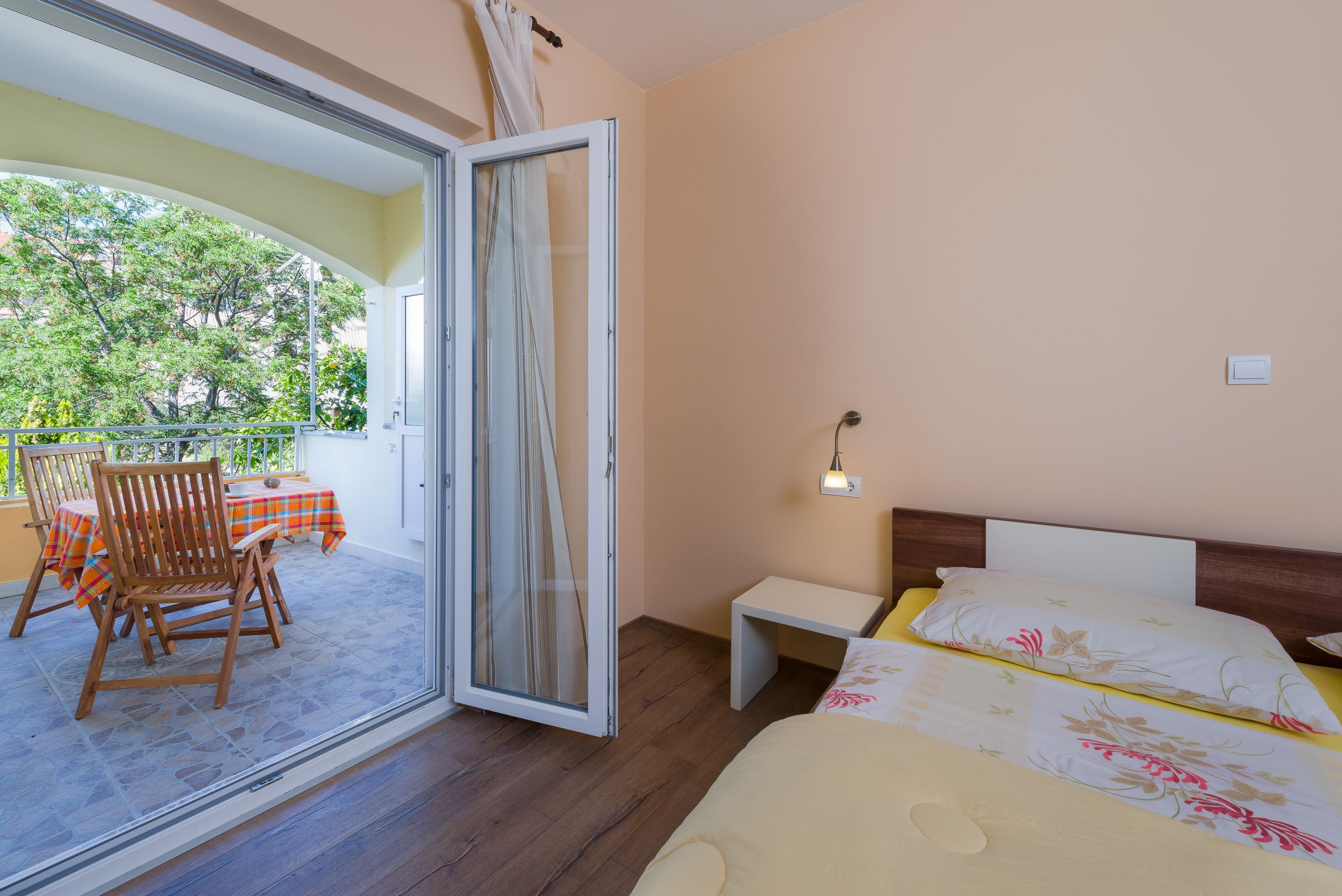 A bedroom with a bed, a nightstand, a chair, a table with a tablecloth, and a balcony with a chair and a table.