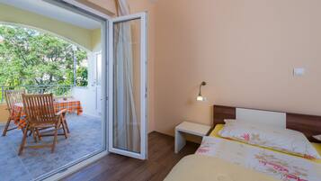 A bedroom with a bed, a nightstand, a chair, a table with a tablecloth, and a balcony with a chair and a table.