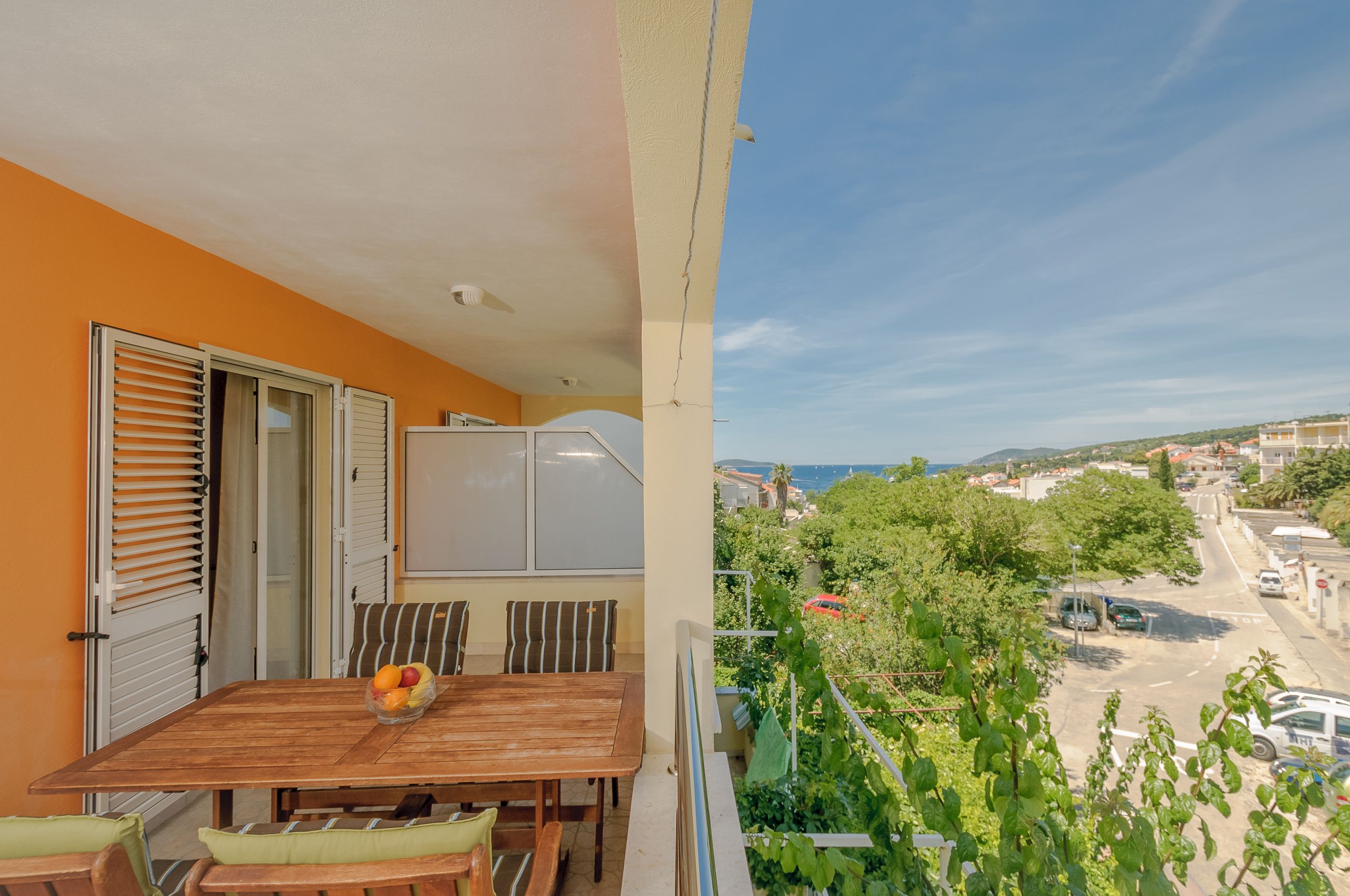 Un balcón con mesa y sillas de madera, un frutero y vistas a un pueblo y al mar.