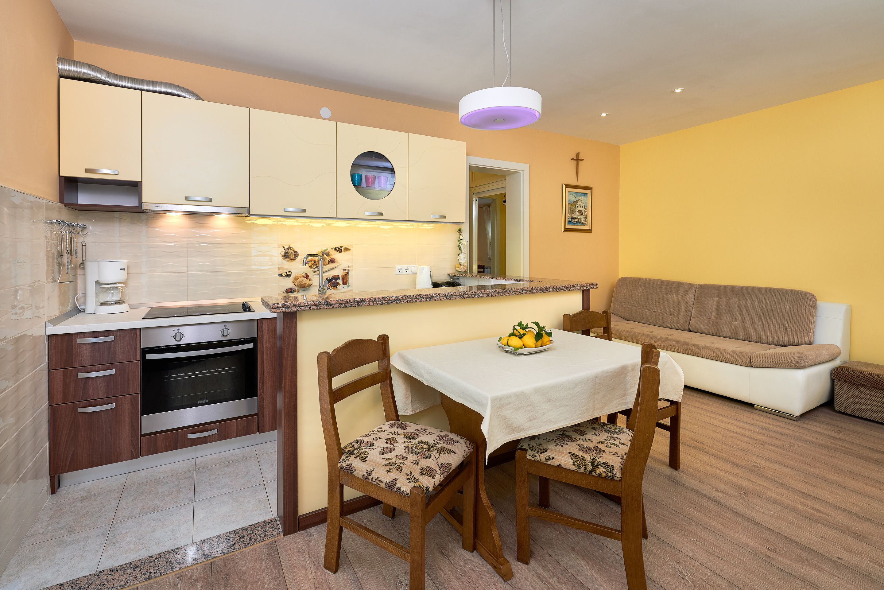 A compact kitchen with modern appliances, a dining area with two chairs, and a sofa in a yellow-walled living space.