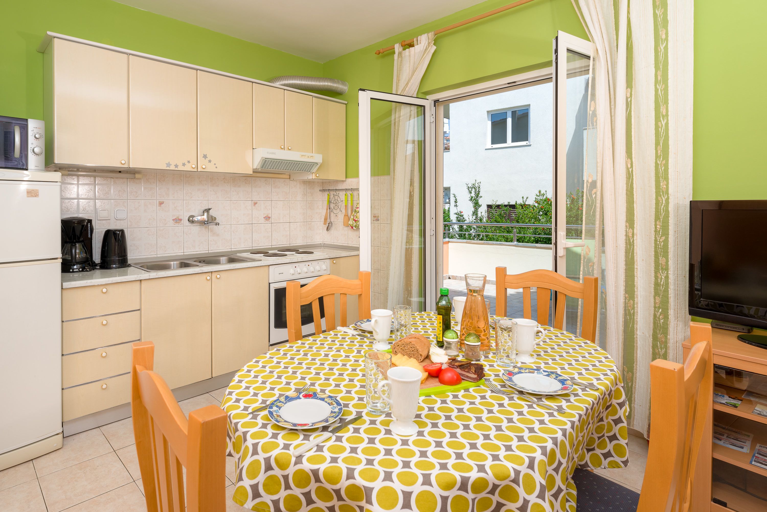 Een kleine keuken met een eethoek, een televisie en uitzicht op een balkon.