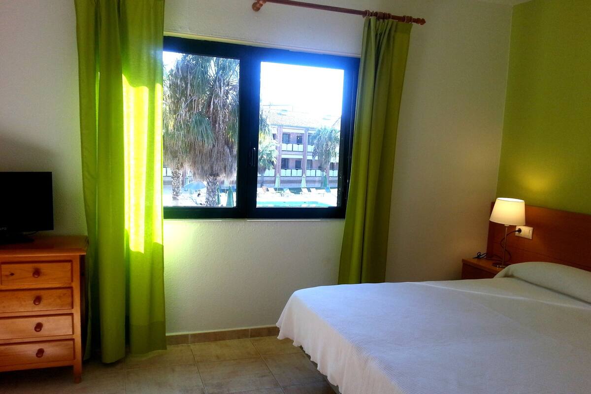 Une chambre d’hôtel avec un lit, une table de chevet avec une lampe, un téléviseur posé sur une commode en bois et une fenêtre avec des rideaux verts.