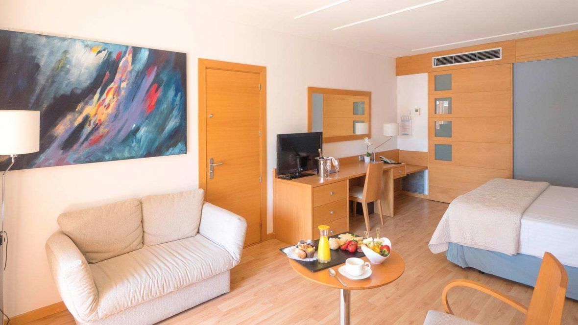 A hotel room with a sofa, a desk with a computer, a bed, and a dining table with breakfast.