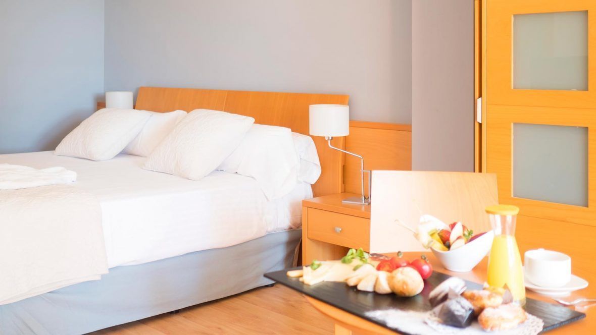 Habitación de hotel con cama, mesita de noche, lámpara y bandeja de desayuno con frutas y pasteles.