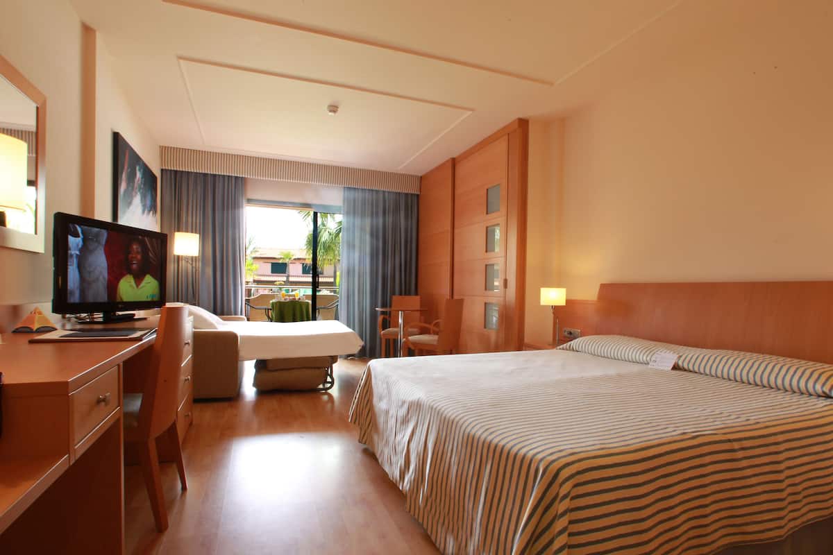 Une chambre d’hôtel comprenant un lit, un bureau avec une télévision, une chaise, une lampe de bureau et une fenêtre avec des rideaux.