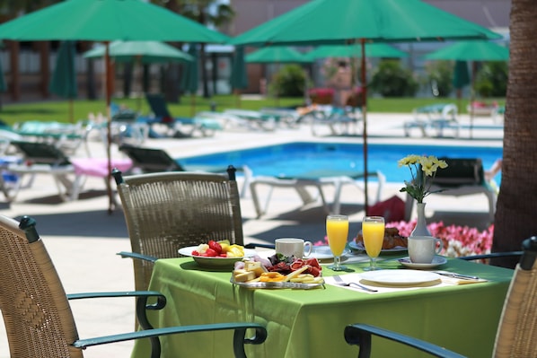 A dining table with breakfast items, a pool in the background, and green umbrellas.