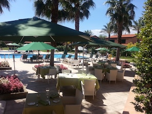 A poolside area with outdoor seating and umbrellas, surrounded by palm trees and a clear sky.