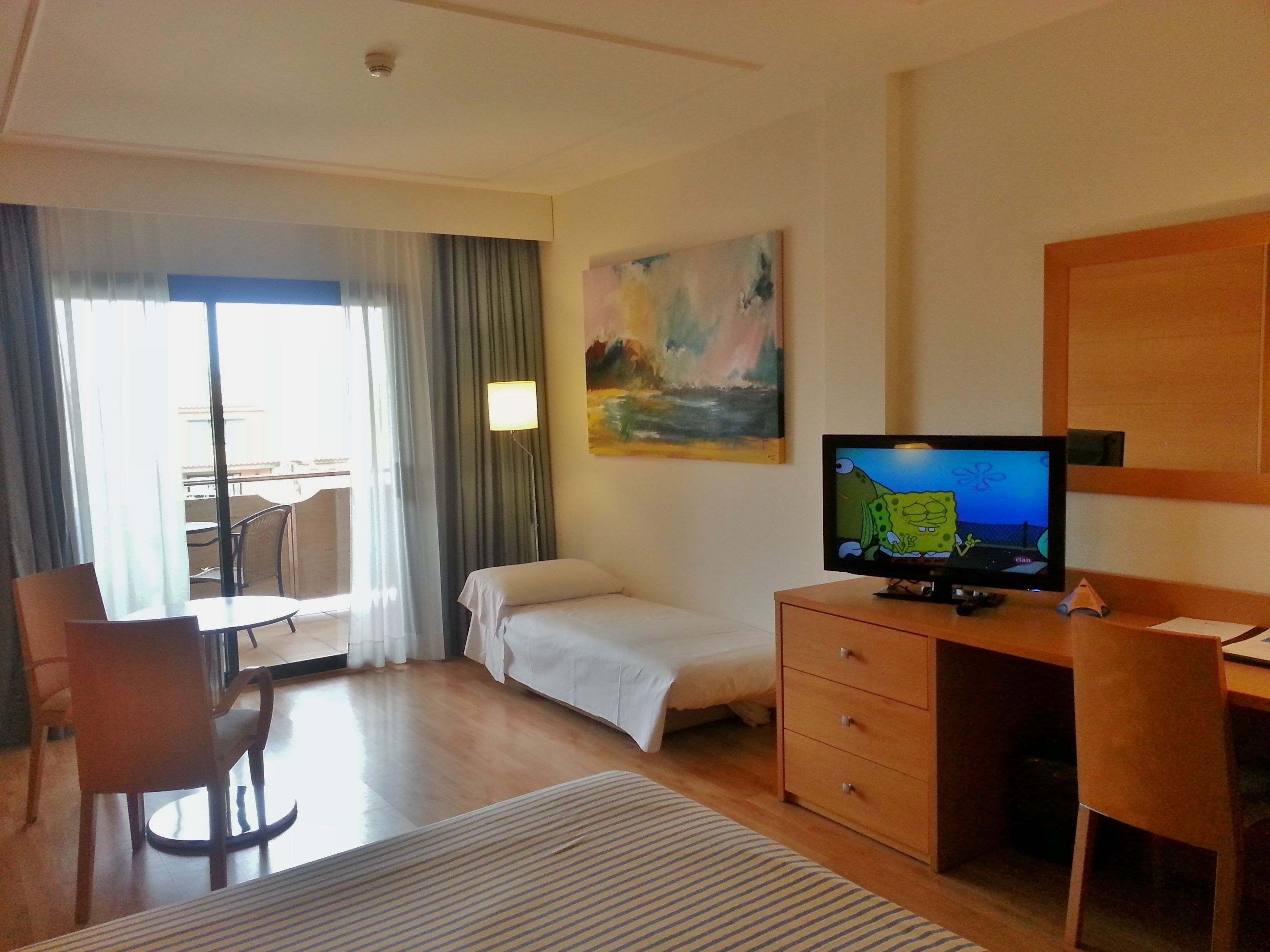 A hotel room with a bed, a desk, a chair, a TV, and a painting on the wall.
