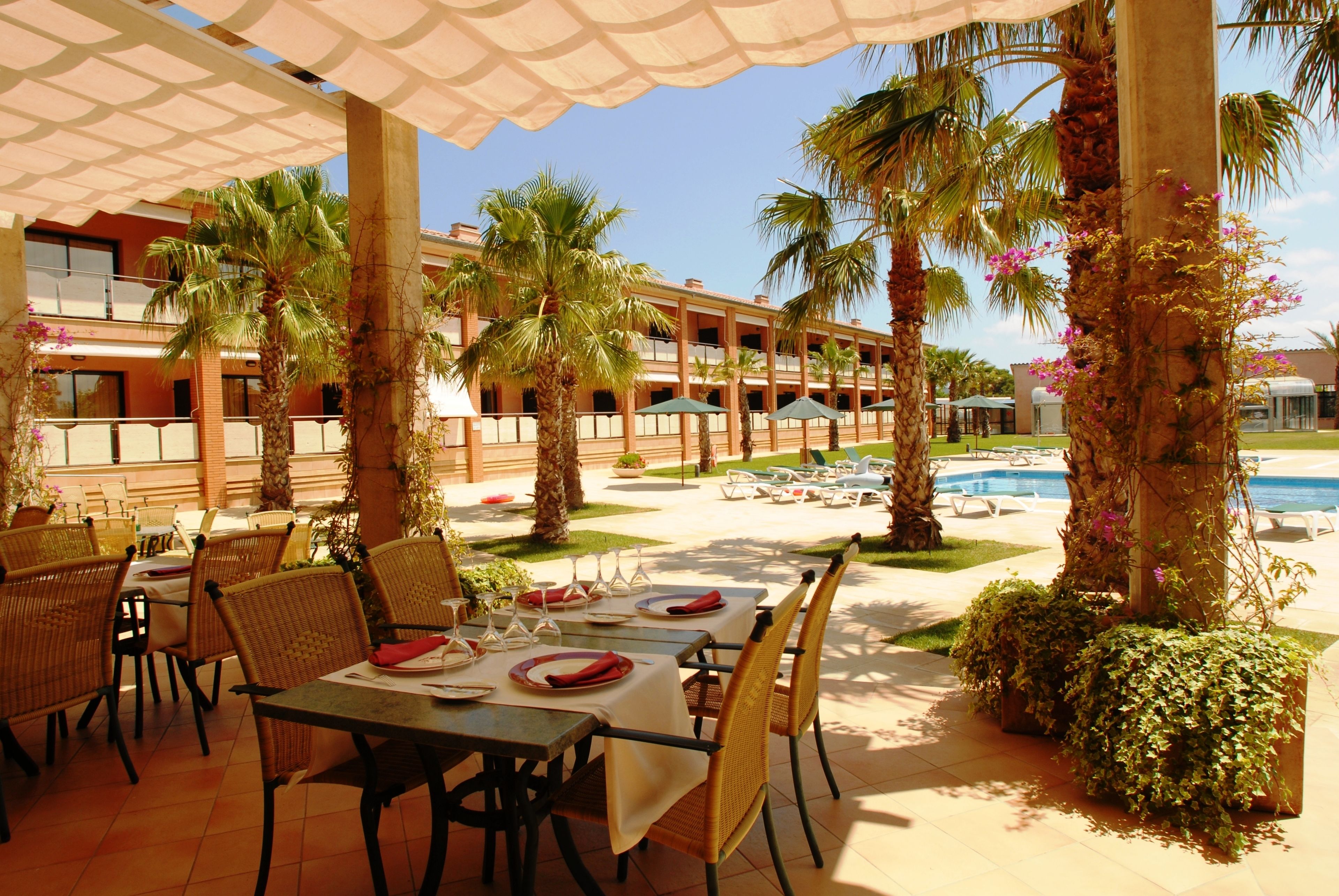 An outdoor dining area with tables and chairs, palm trees, and a pool in the background.