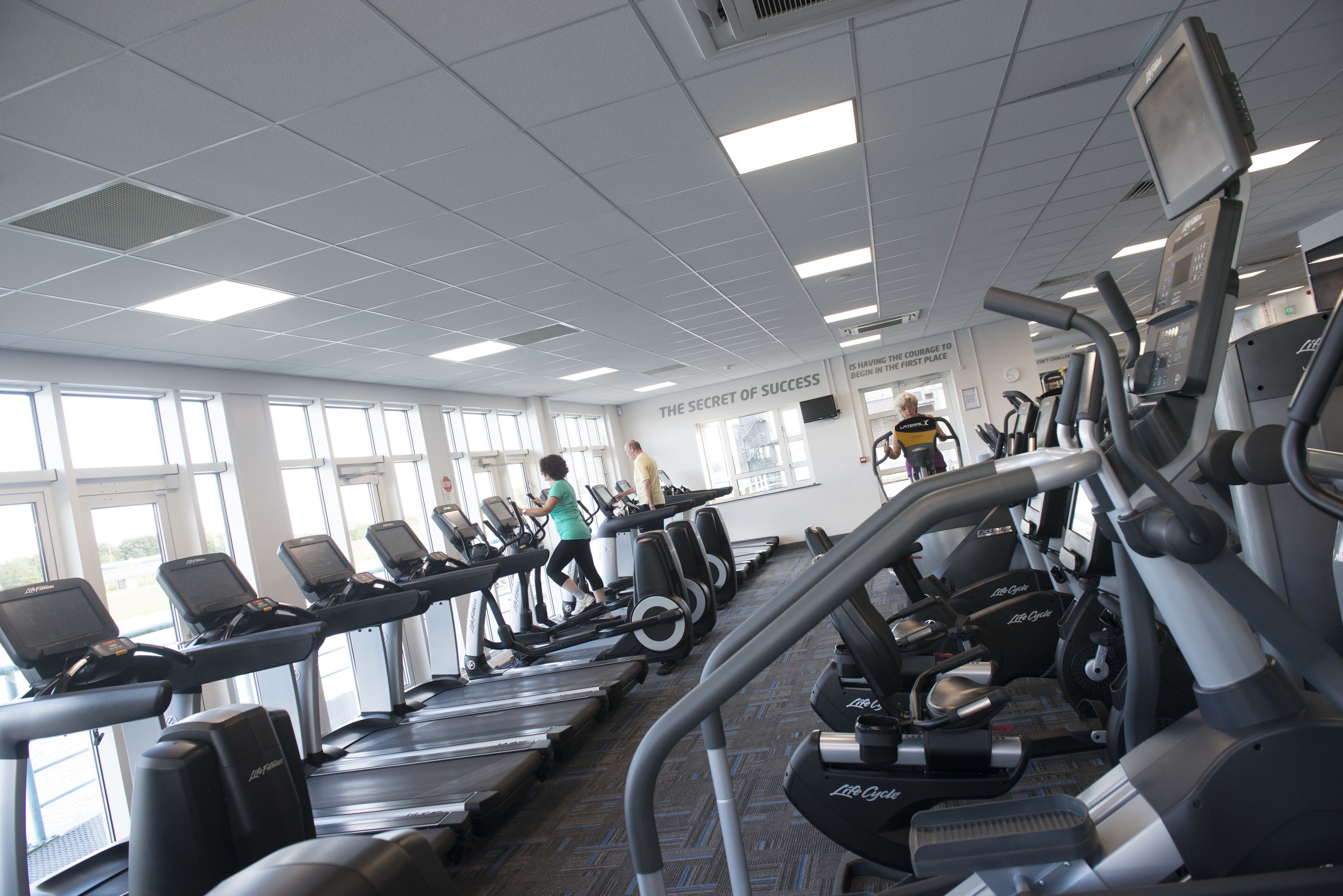 Ein Fitnessraum mit Laufbändern und Crosstrainern, großen Fenstern und einem motivierenden Spruch an der Wand.