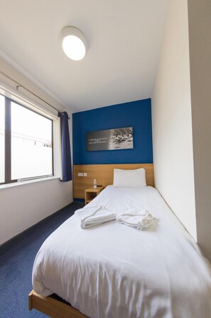 Une chambre d’hôtel avec un lit simple, une table de chevet, un tableau accroché au mur, une fenêtre avec des rideaux et une moquette bleue.