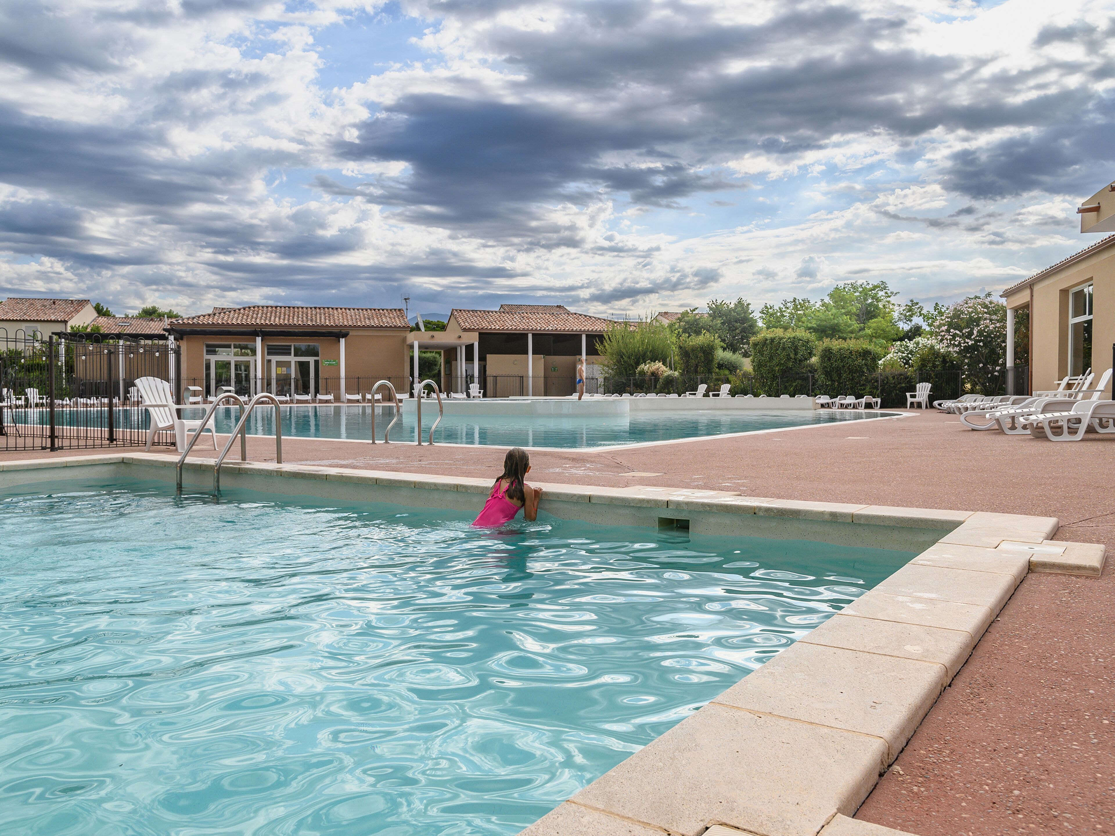 A swimming pool with a child in the water, surrounded by lounge chairs and a covered area with a tiled roof.