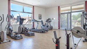 Un gimnasio amplio con diversa maquinaria de cardio y entrenamiento de fuerza, ventanas grandes y piso de madera.