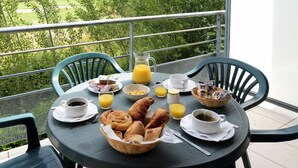 Mesa de desayuno con cruasanes, jugo, café y una cesta de pan en un balcón con vistas a árboles.