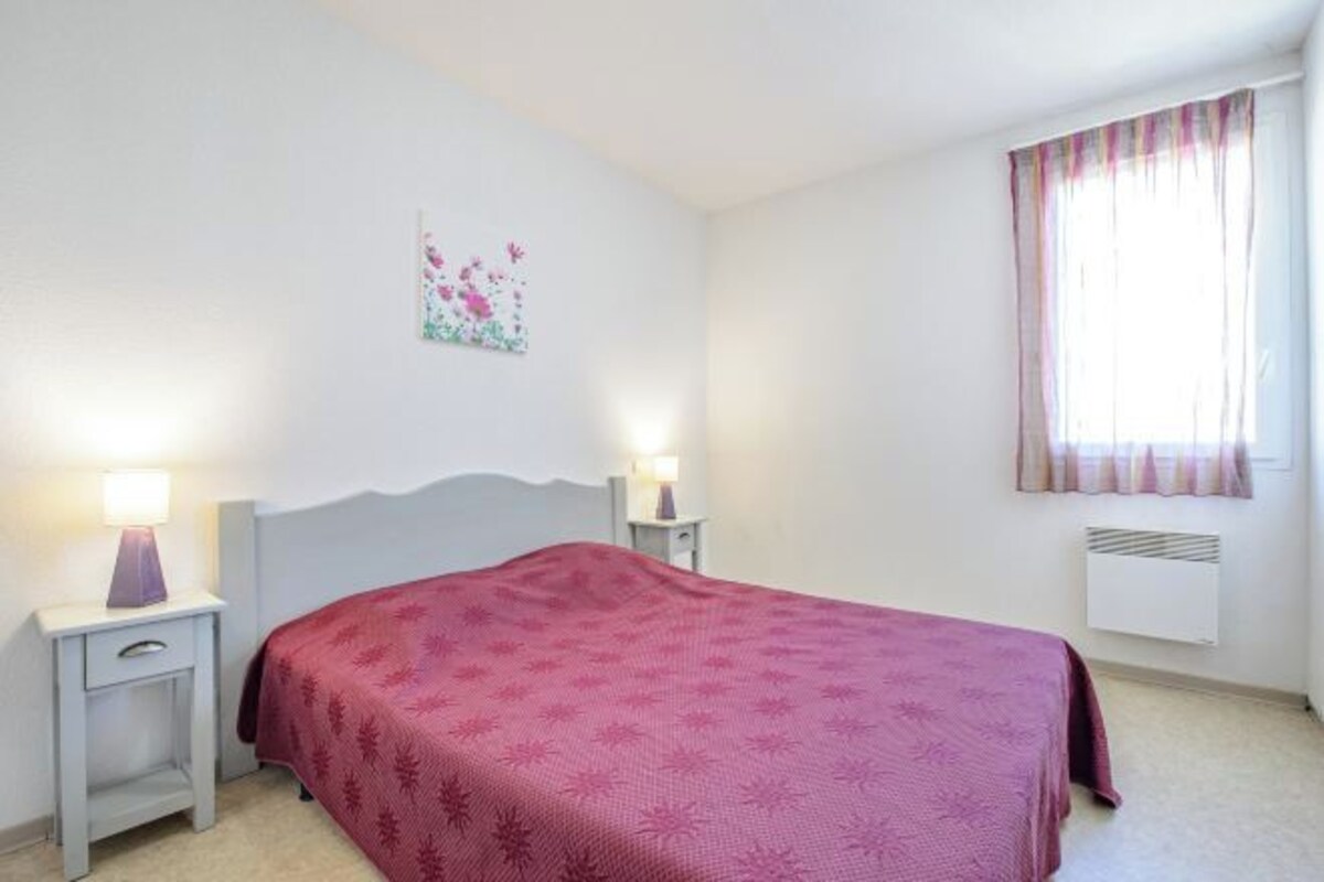 A bedroom with a bed, two bedside tables with lamps, a window with curtains, and a wall-mounted air conditioner.