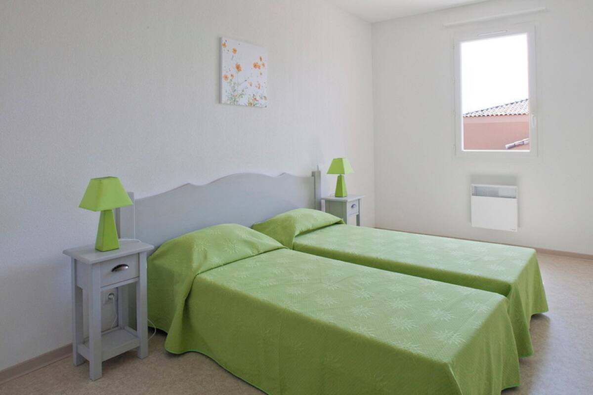 A room with two single beds, a nightstand with a lamp, a small table, and a window with a view of a building.